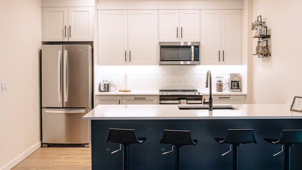 A modern kitchen with white cabinets, stainless steel appliances, a central island with bar stools, and a microwave above the oven. - Revy To Go - Unique 2-Bedroom Condo (Revelstoke)