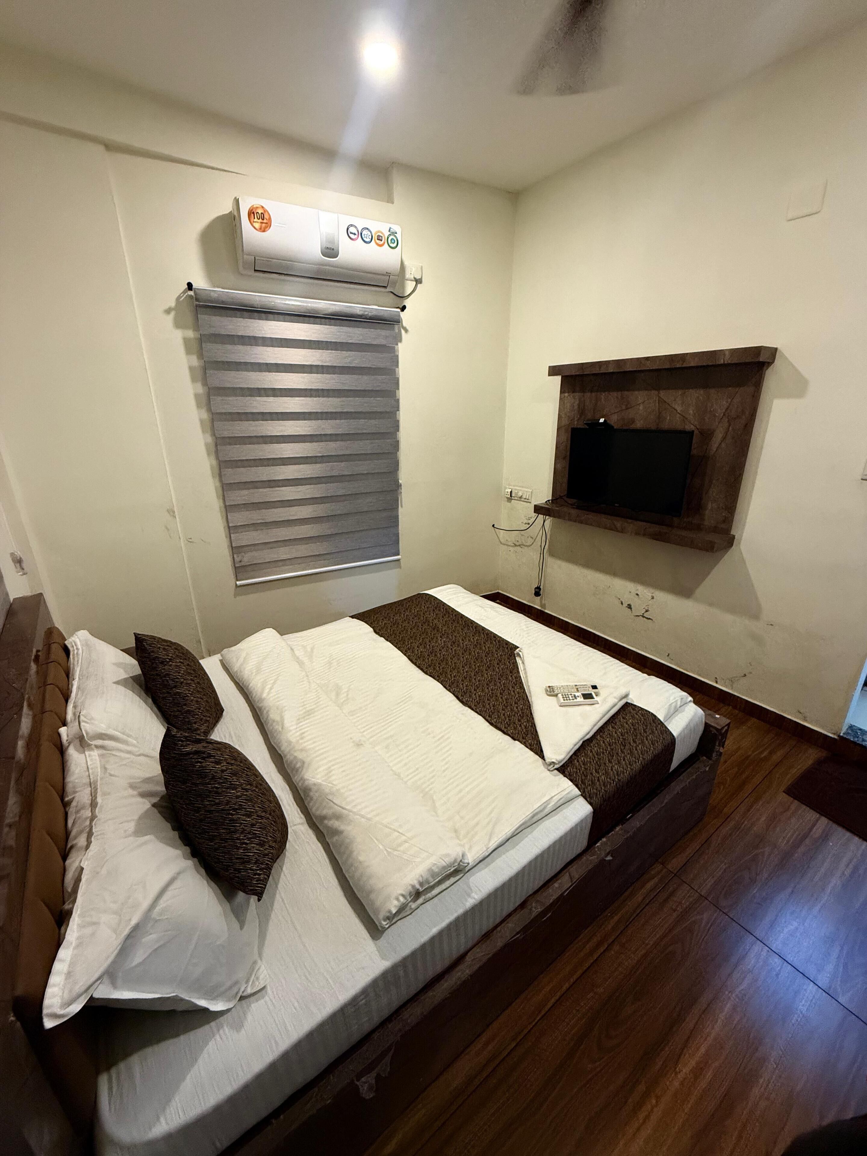 Una cama bien hecha con un control remoto, un televisor montado en la pared y un ventilador de techo en una habitación con suelo de madera y una ventana con persianas.