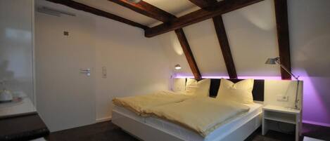 A bedroom with a bed, bedside tables, a nightstand, a ceiling light fixture, and exposed wooden beams.