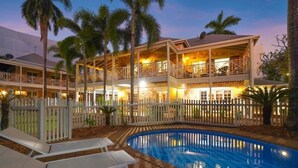 Um hotel de dois andares com piscina, palmeiras e espreguiçadeiras.