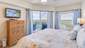 Un dormitorio con una cama grande, televisor, cómoda de mimbre y un balcón con vista al océano.