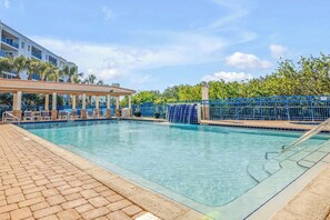 A swimming pool with a tiled deck, a covered seating area, and a blue fence.