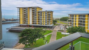 Un immeuble jaune à plusieurs étages, doté de balcons, avec une marina en arrière-plan et une zone herbeuse parsemée de sentiers.