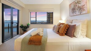 Un dormitorio amplio con una cama grande, vista al mar a través de una puerta corrediza y un cuadro de palmeras en la pared.