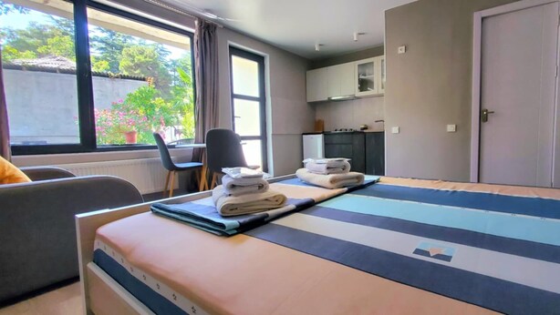 Une chambre d’hôtel équipée d’un lit, d’un bureau, d’une chaise, d’une petite kitchenette et d’une fenêtre donnant sur un espace verdoyant.