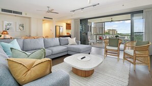 Ein modernes Wohnzimmer mit einem grauen Ecksofa, einem runden Couchtisch, Korbstühlen und einer großen Schiebeglastür, die zu einem Balkon mit Stadtblick führt.