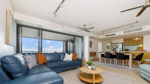 Ein modernes Wohnzimmer mit einem blauen Ledersofa, einem runden Holztisch und Blick auf den Himmel durch große Fenster.
