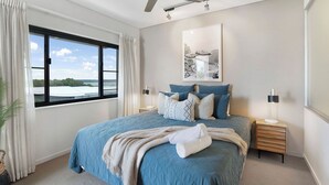 Een slaapkamer met een groot bed, een plafondventilator, een raam met gordijnen, een nachtkastje met een lamp en een lijst met een afbeelding aan de muur.