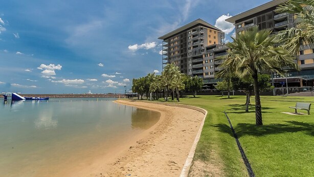 A beachfront hotel with a sandy beach, palm trees, and a clear sky. - Enchanting Waterfront Lagoon Views + Restaurants (Darwin)