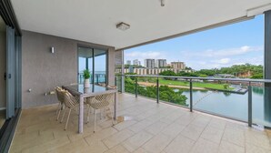 A balcony with a view of a river and buildings, a table with chairs, and a potted plant. - Enchanting Waterfront Lagoon Views + Restaurants (Darwin)