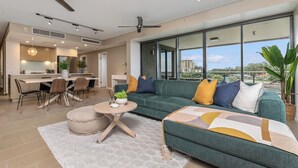 A modern living room with a green sofa, a round coffee table, and a dining area with a kitchen in the background. - Enchanting Waterfront Lagoon Views + Restaurants (Darwin)