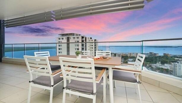 A balcony with a dining table and chairs, overlooking a cityscape and the sea during sunset.