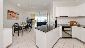 Cozinha moderna com armários brancos, bancada preta e forno embutido. Há uma área de refeições com mesa e cadeiras, e um ventilador de teto.