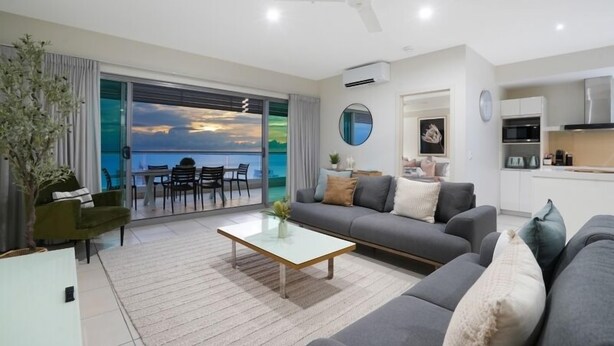 Ett modernt vardagsrum med en grå soffa, ett soffbord i glas och utsikt  över havet genom stora fönster.