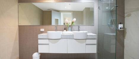 A modern bathroom with a large mirror, two sinks, a glass shower enclosure, and a trash bin.
