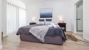 A bedroom with a large bed, a ceiling fan, a nightstand with a lamp, and a painting of the ocean on the wall. - Scenic CBD Getaway, Harbour Views, Pool/Gym, Cafes (Darwin)