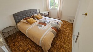 Un dormitorio con un cabecero capitonado, una cama con ropa de cama estampada, una mesita redonda pequeña, una ventana con cortinas y un radiador.