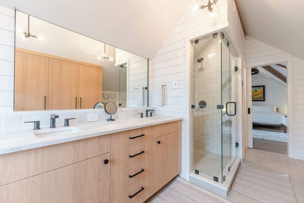 A modern bathroom with a large mirror, wooden cabinets, a glass shower enclosure, and a visible bedroom with a bed.