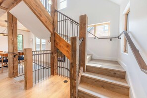 Un escalier en bois avec des rampes en métal, un coin repas avec des tables et des chaises, et de grandes fenêtres laissant entrer la lumière naturelle.