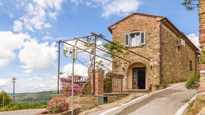 A stone building with a sloped roof, a wooden pergola with climbing plants, and a paved road leading to it.