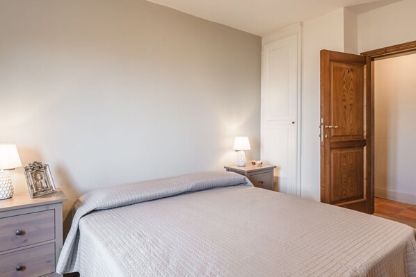 Un dormitorio con una cama, dos mesitas de noche, una mesita auxiliar, una puerta de madera y una pared blanca.