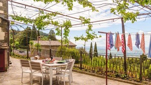 A stone patio with a table and chairs, a pergola with climbing plants, and a view of the sea.