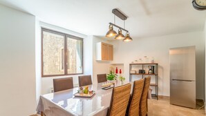Een eetgedeelte met een tafel gedekt voor vier personen, een keuken met een koelkast, en een raam met uitzicht op een stenen muur.