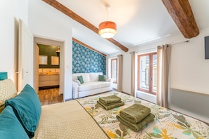 A bedroom with a bed, a sofa, a wooden ceiling beam, a hanging light fixture, and a window with curtains. - Holiday Home ‘La Bonbonnière’ – Charm and Comfort at the Foot of Mont Ventoux (Malaucène)