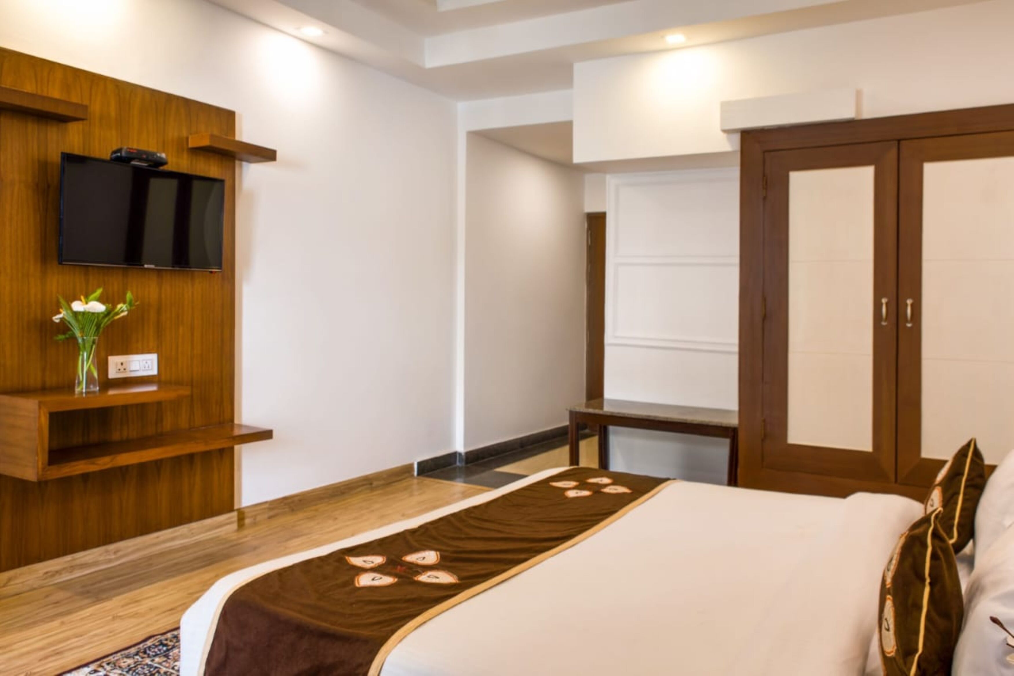 A hotel room with a bed, a TV mounted on the wall, a wooden wardrobe, and a small table with a vase of flowers.