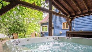 Ein Whirlpoolbereich mit einer hölzernen Pergola darüber, ein blaues Haus im Hintergrund und eine Terrasse mit Gartenmöbeln.