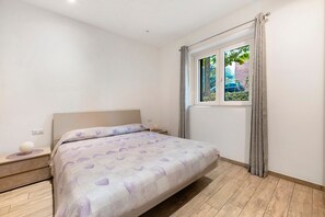 A bedroom with a bed, a nightstand, a window with curtains, and a view of greenery outside.