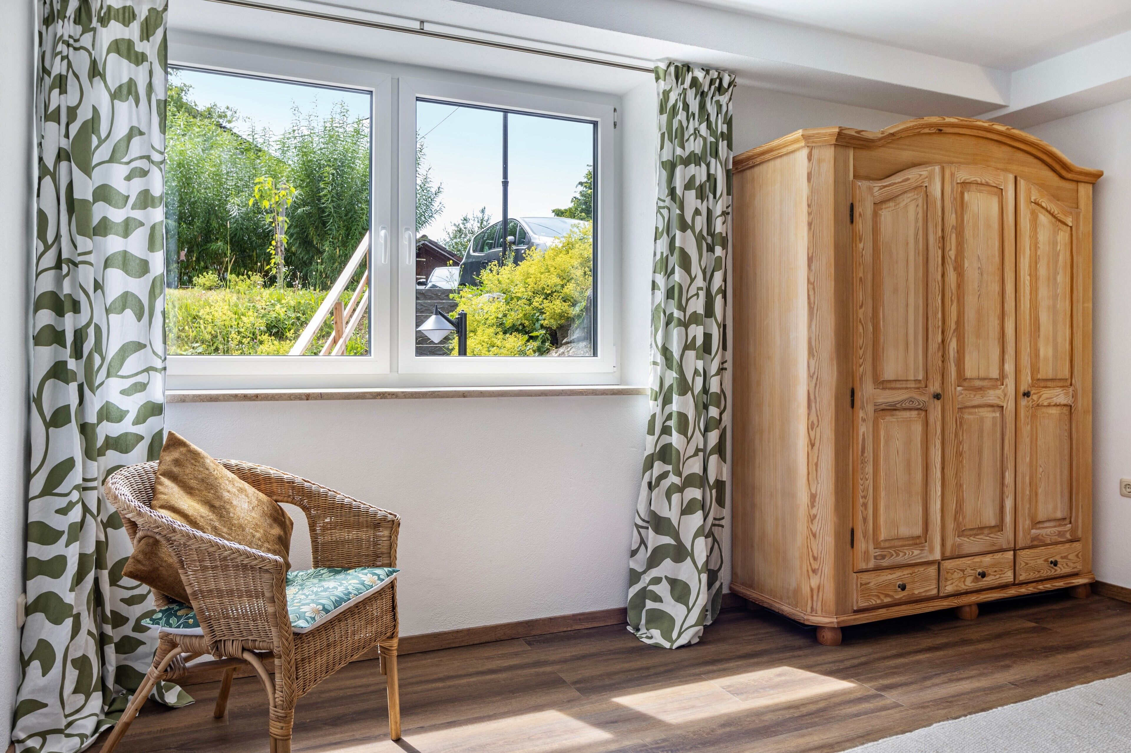 Ein Zimmer mit großem Fenster, einem Korbstuhl und einem Holzschrank.