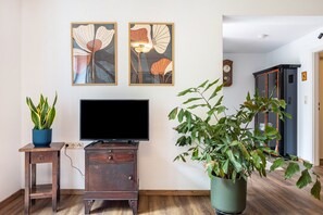 A living room with a flat-screen TV, wooden furniture, two framed floral paintings, a potted plant, and a clock on the wall. - Apartment 'Krottenhill 2 ¼' with Sauna, Garden, and Wi-Fi (Ingenried)