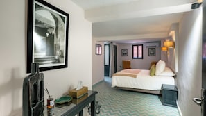 Una habitación de hotel con cama, una mesita de noche con caja de pañuelos, un cuadro en la pared y una ventana con cortinas.
