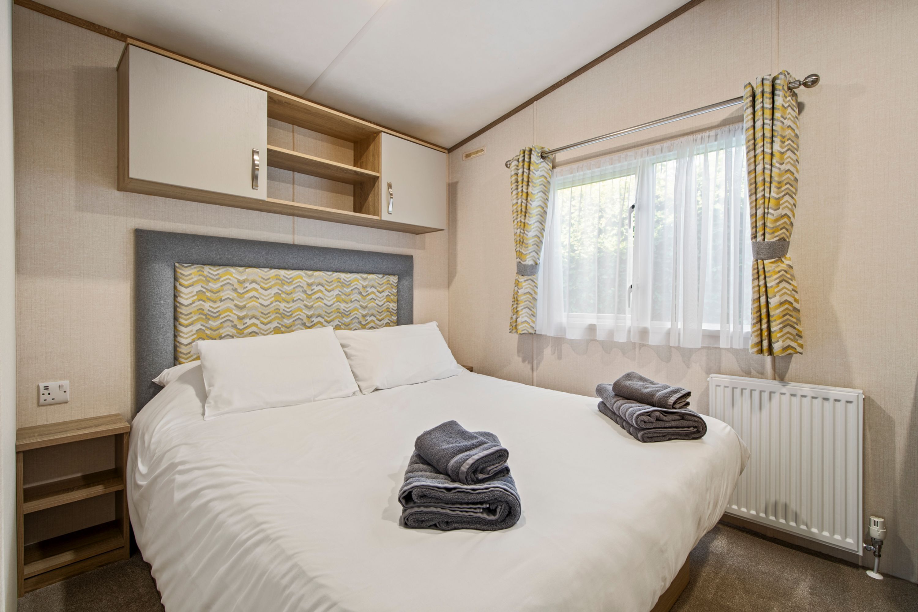 Ein ordentlich bezogenes Bett mit zwei gefalteten Handtüchern, einem Kopfteil mit Zickzackmuster, einer eingebauten Ablage über dem Bett und einem Fenster mit Vorhängen.