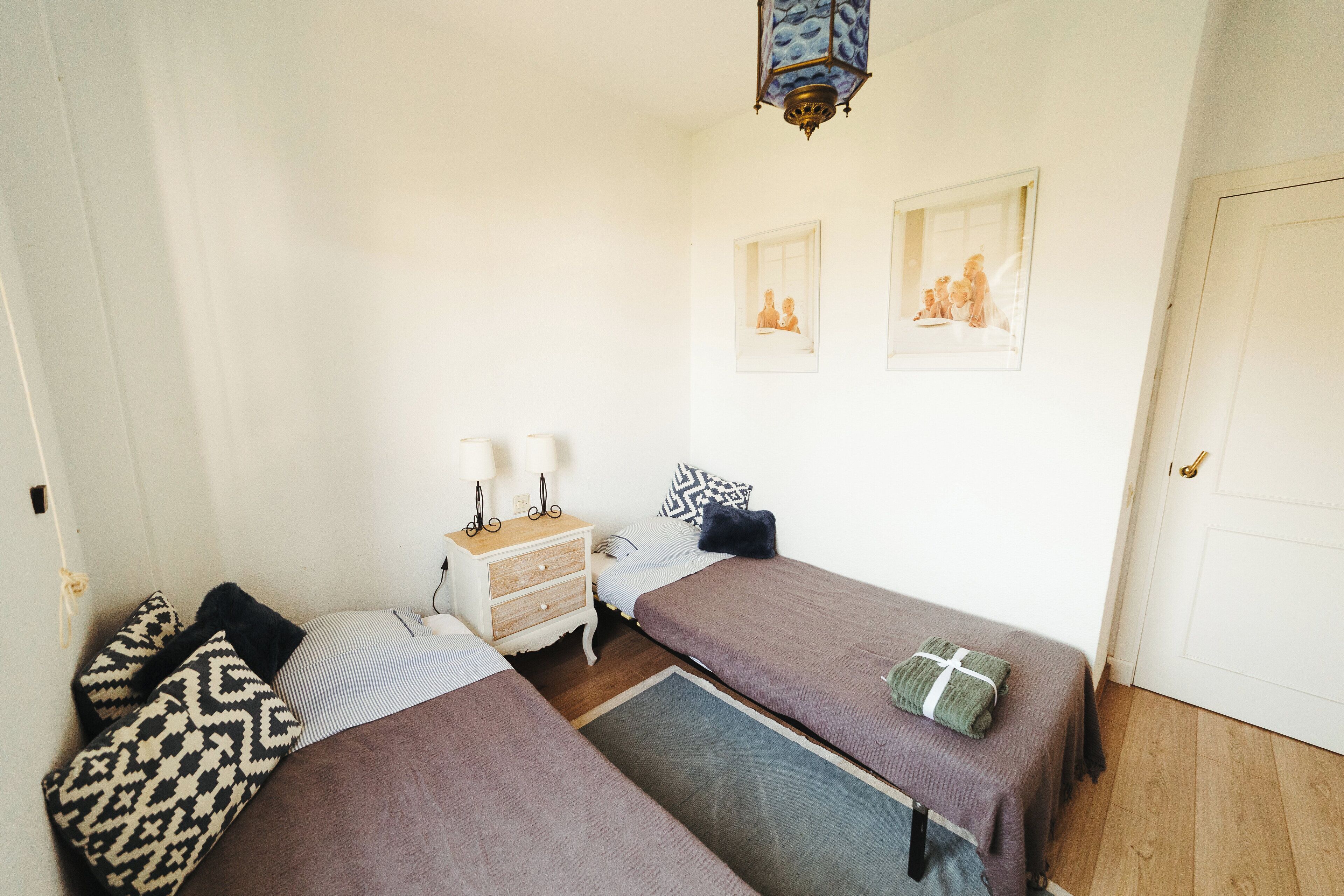 Une petite pièce bien éclairée, avec deux lits, une table de chevet et une lampe fixée au mur.