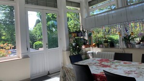 Et spiseområde med et bord dækket af en blomstret dug, omgivet af stole. Store vinduer med persienner, og en radiator er synlig på væggen.