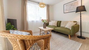 Ett vardagsrum med en grön soffa, flätade fåtöljer, ett litet bord med en vas, en golvlampa och ett stort fönster med gardiner.