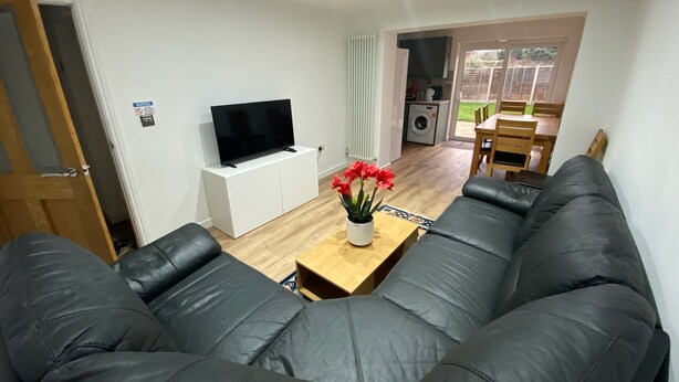 Een woonkamer met een bank, een televisie op een wit kastje, een bijzettafeltje met bloemen, en een keuken op de achtergrond.