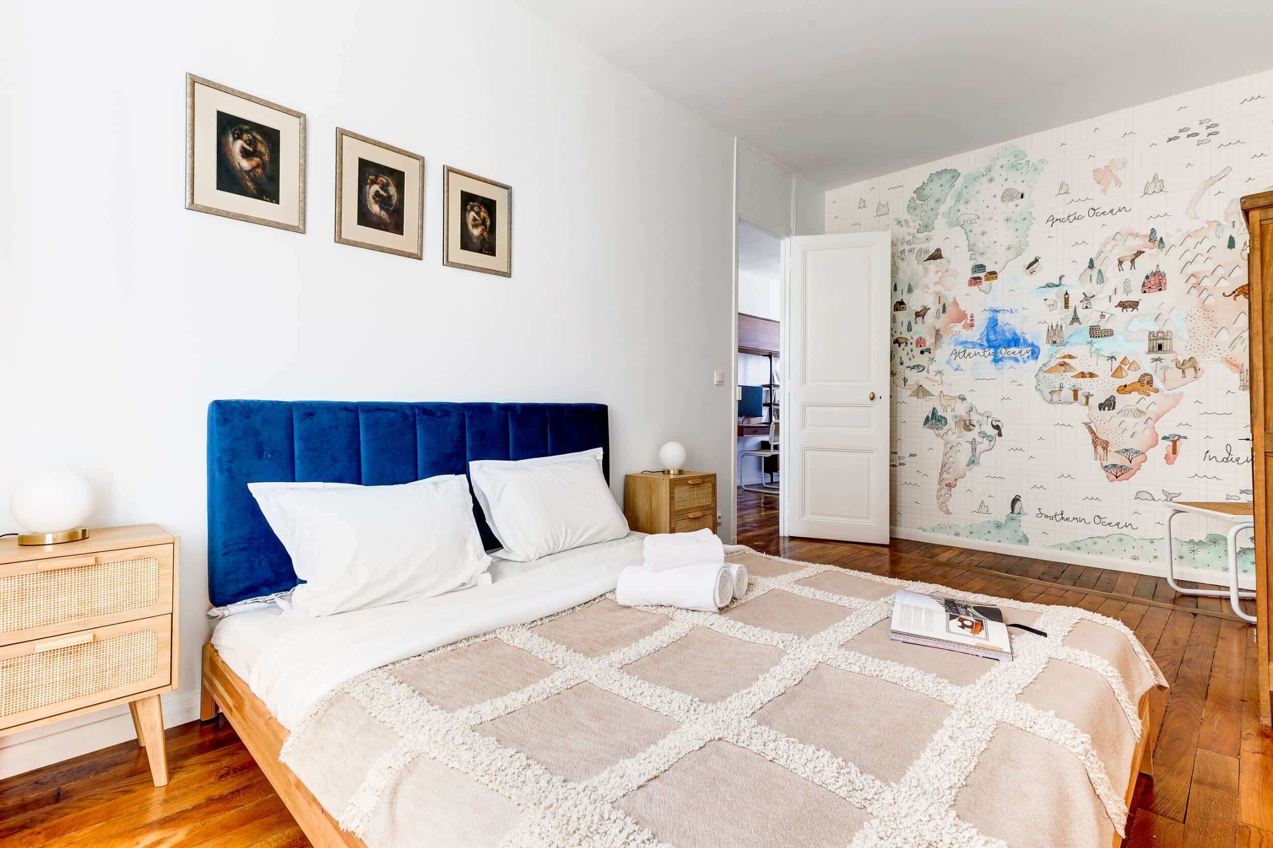 Un dormitorio con cabecera azul, ropa de cama blanca, una mesita de noche de ratán, una silla blanca y un mural que representa diversas escenas marinas.