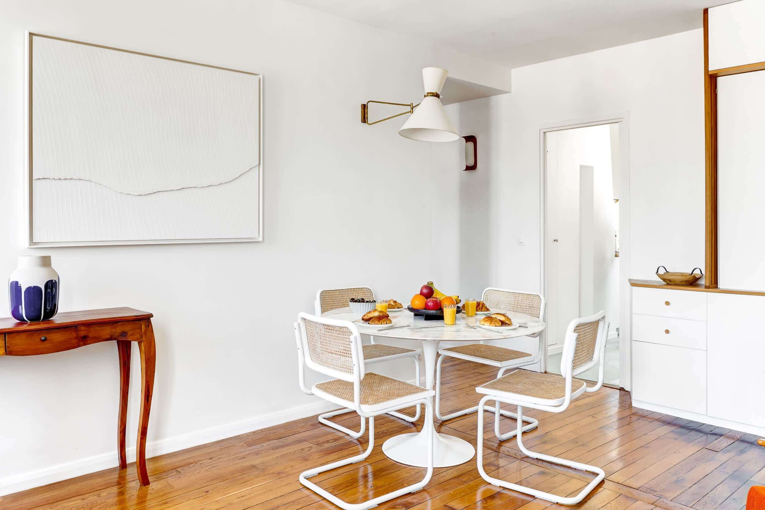 Un coin repas avec une table ronde, des chaises blanches et un saladier rempli de fruits.
