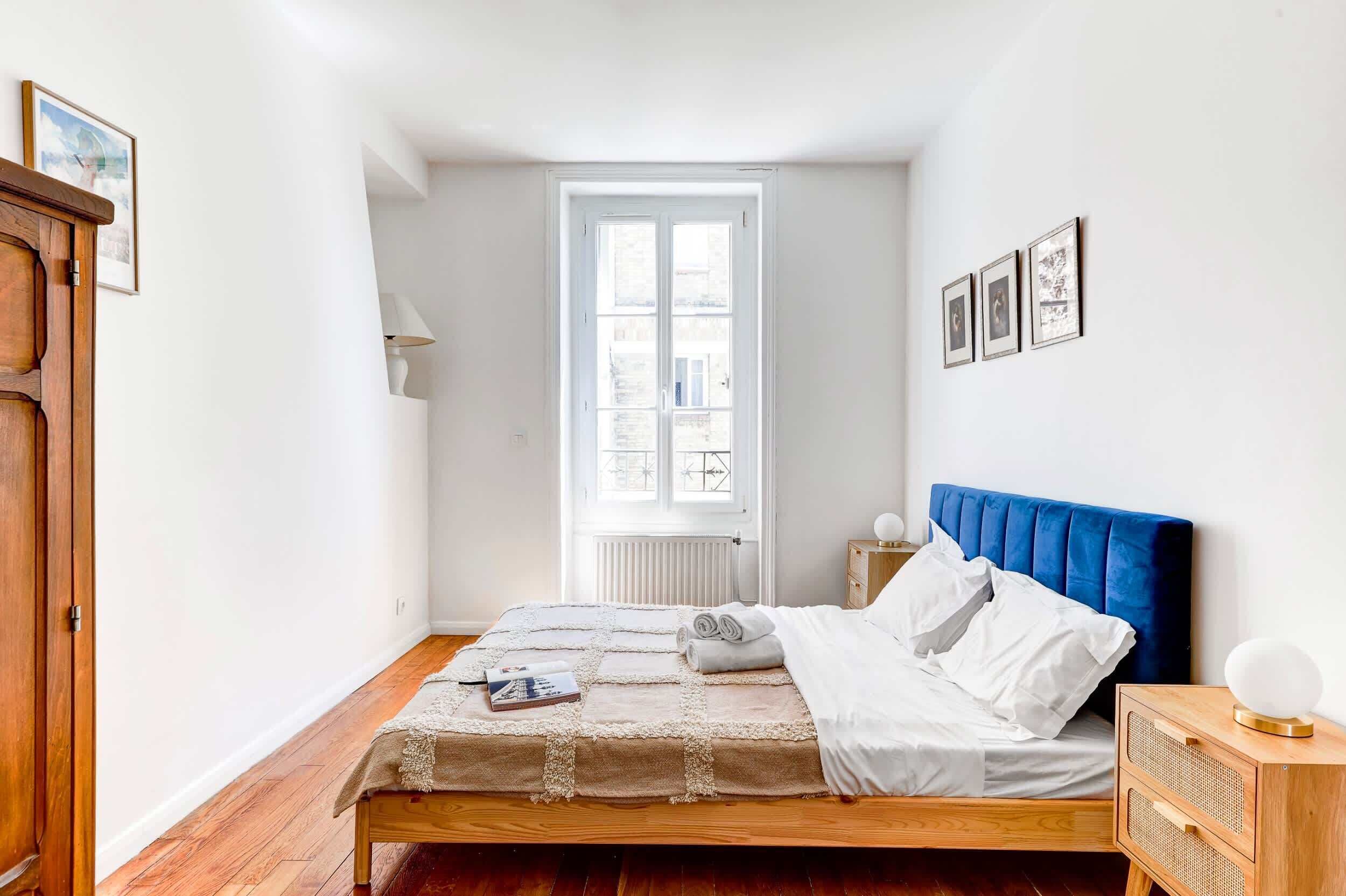 Een slaapkamer met een houten bed, witte beddengoed, een blauw hoofdeinde, een nachtkastje met een lamp, en een raam met uitzicht op gebouwen.