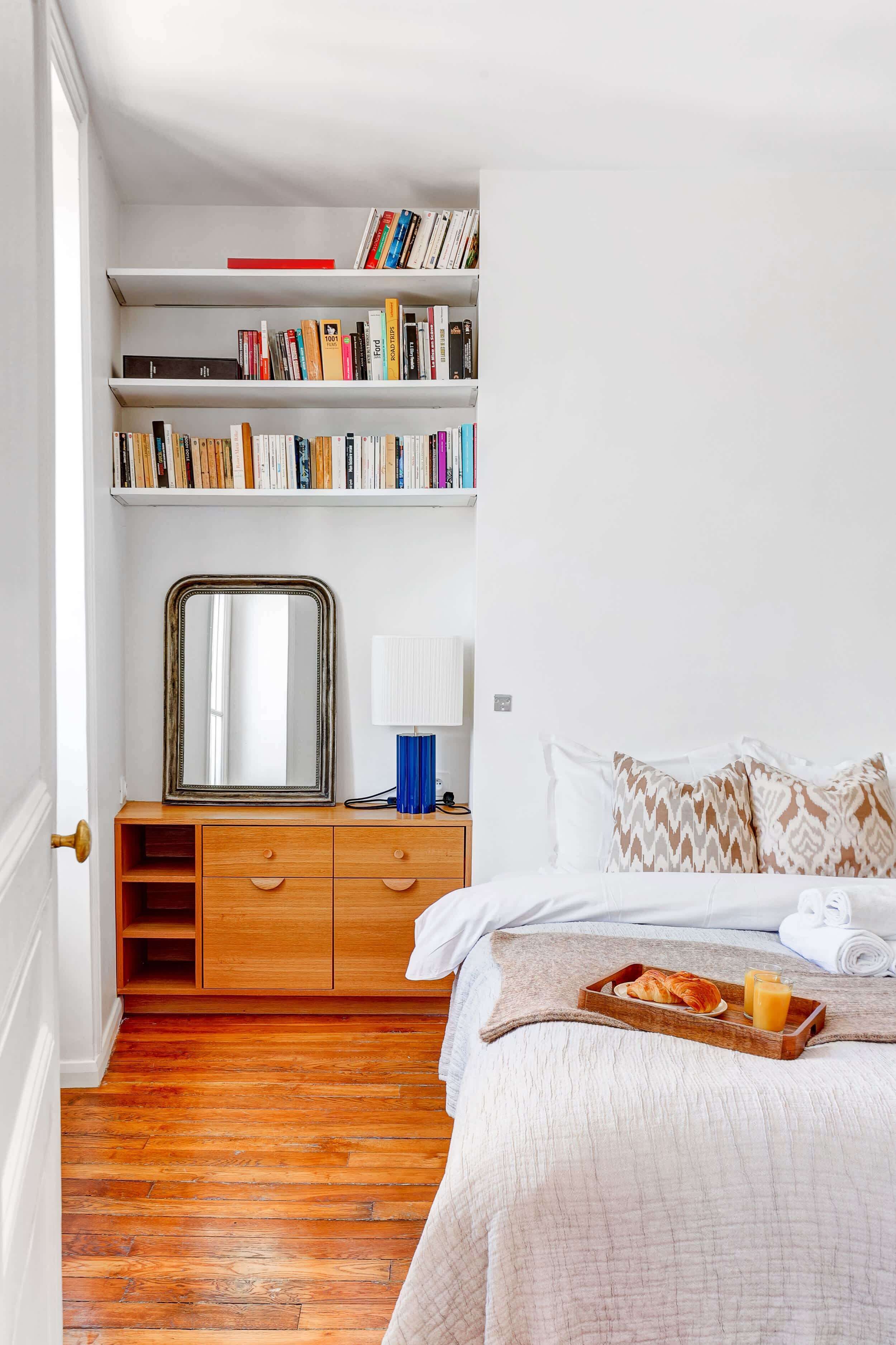 A bedroom with a wooden dresser, a mirror, a bedside table with a lamp, a bed with pillows, a tray with croissants and juice, and a bookshelf with books.
