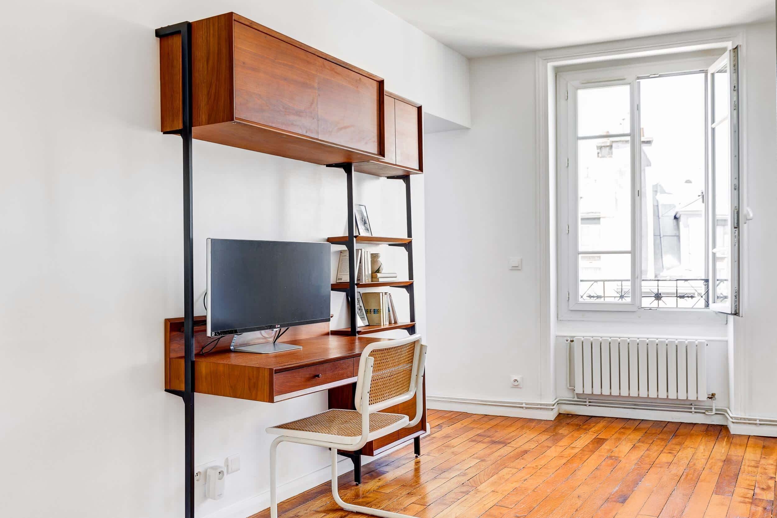 A modern room with a wooden desk, a television, a bookshelf, and a window with a view of buildings.