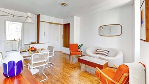 Un salon moderne avec un canapé blanc, des fauteuils orange et un coin repas comprenant une table et des chaises en bois.