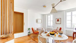 Un espace de vie lumineux et ouvert, doté d’un plancher en bois, de murs blancs et d’une table à manger ronde entourée de chaises. Une fenêtre laisse entrer la lumière naturelle et un ventilateur de plafond est installé.