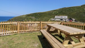 Une table et un banc de pique-nique en bois se trouvent dans un espace verdoyant, délimité par une clôture en bois, et offrent une vue sur un paysage côtier avec une maison et des véhicules.