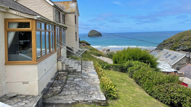 Un bâtiment situé sur la côte, doté de grandes fenêtres donnant sur la mer, un chemin de pierre menant à l’entrée, et une vue sur l’océan et une île lointaine.