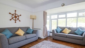 Un salon avec deux canapés gris, un tapis à motifs, un élément de décoration représentant un gouvernail de bateau, une lampe sur pied et de grandes fenêtres avec des rideaux.