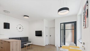 A modern living room with a grey sofa, a wooden dining table with two chairs, a TV mounted on the wall, and a clock. - Beautiful apartment for 3 guests with WIFI, TV and terrace (Železný Brod)